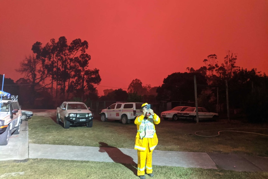 A CFA volunteer takes a photograph on their phone. Behind them the sky glows red.