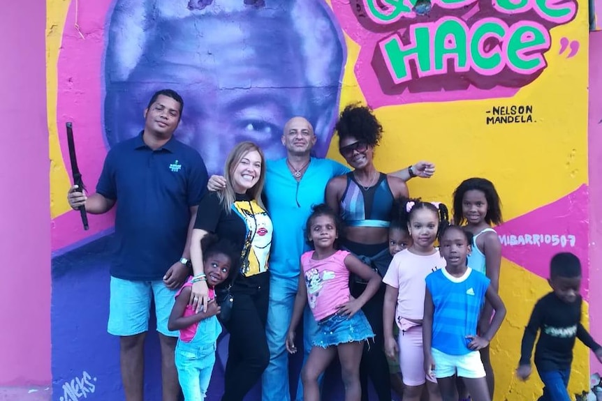 Adults involved in Mi Barrio 507 project stand in front of a colourful mural of Nelson Mendela alongside young children.