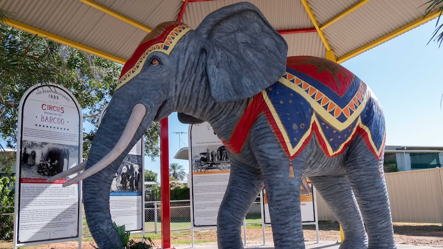A painted lifesize elephant statue stands underneath a colourful rotunda in a park.