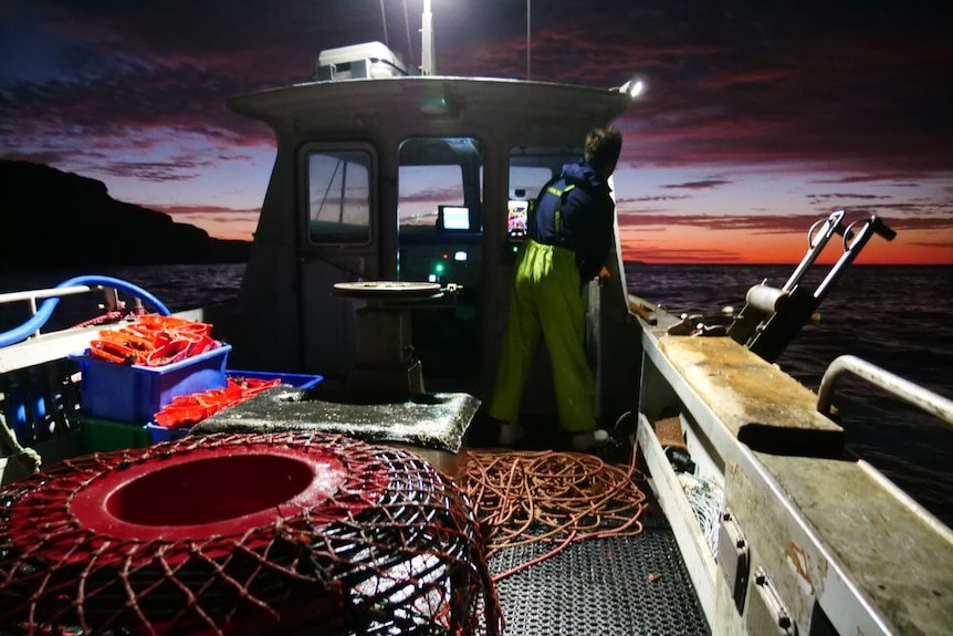 A man with lobster nets on a boat with colourful dawn sky on horizon.