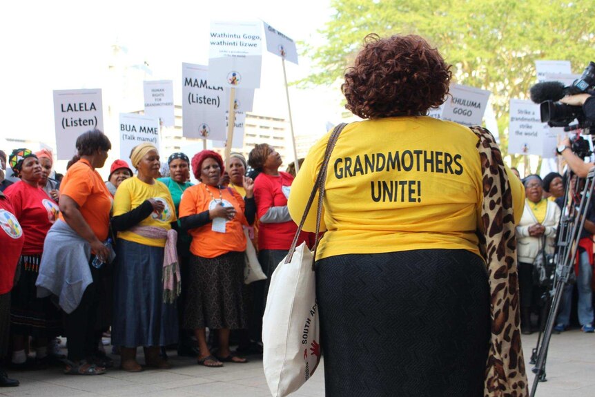 A woman in a yellow t-shirt reading 'Grandmothers Unite' faces a group of women holding signs.