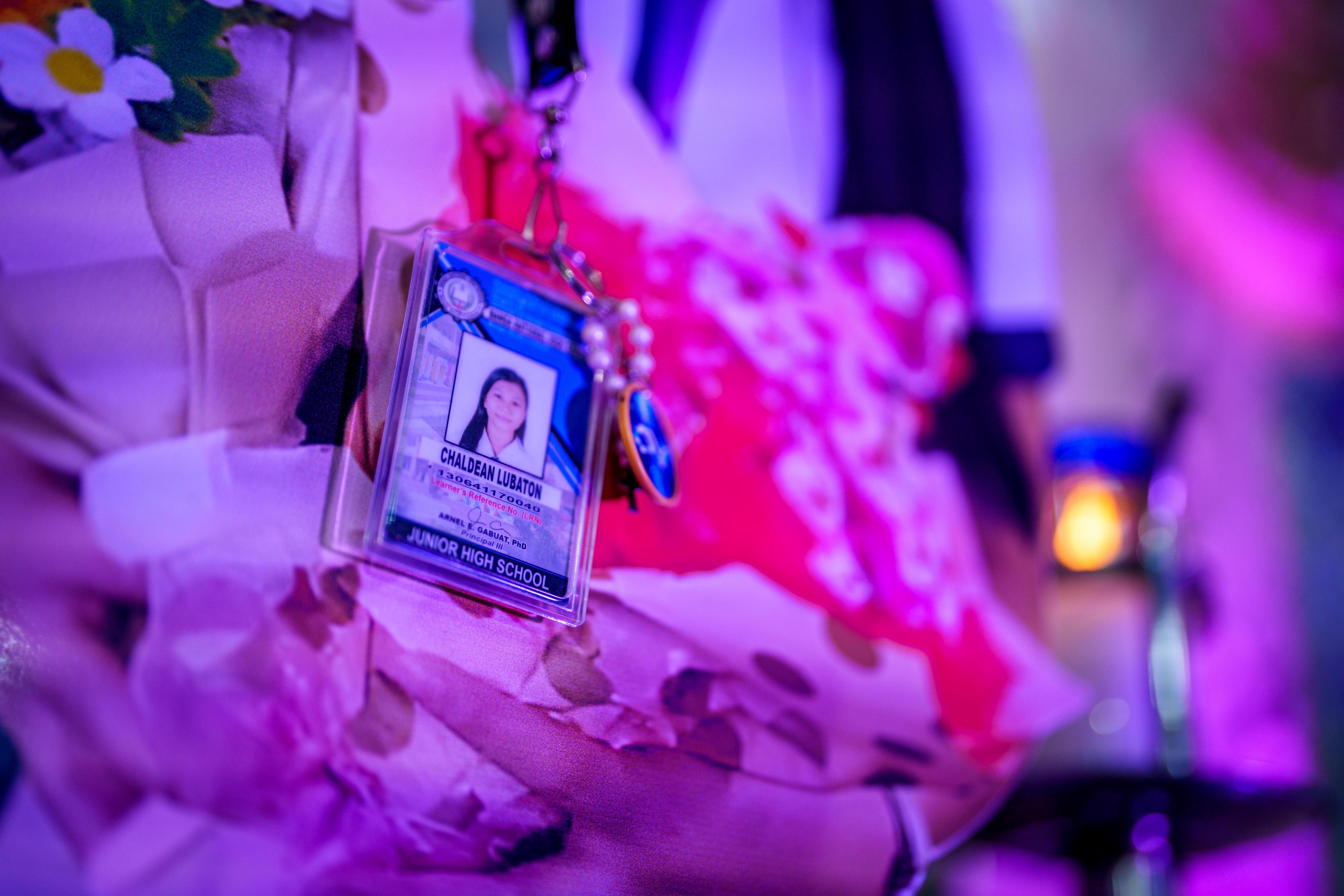 A school identification card belonging to Chaldean Lubaton rests next to a white coffin bathed in purple light.