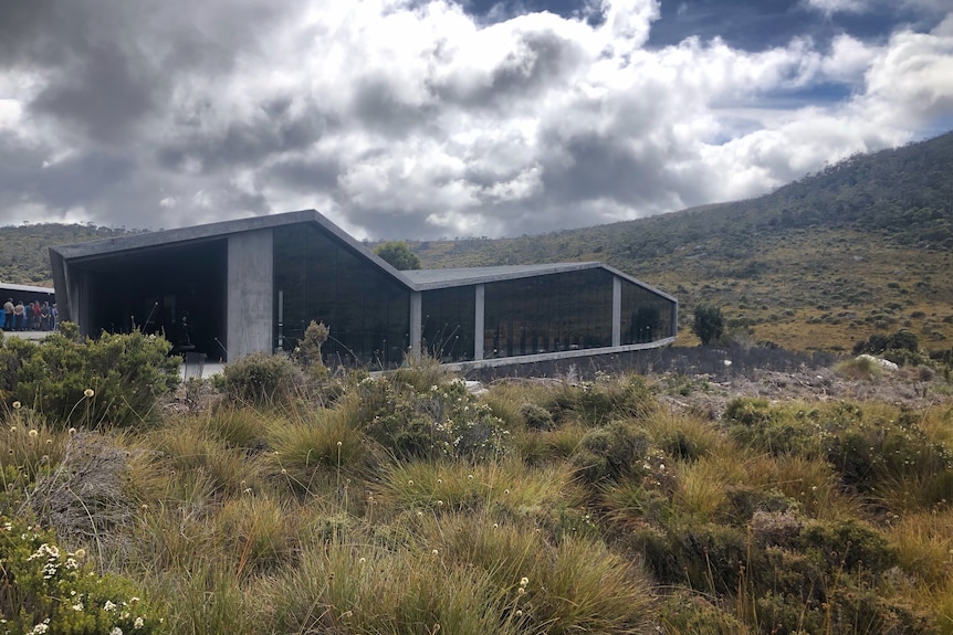 A low-set, concrete building is visible behind small shrubs in an alpine landscape.