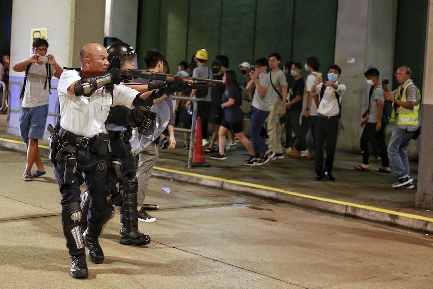 A police officer with an aggressive expression stands on a road in front of a crowd of protesters, pointing a shotgun at them