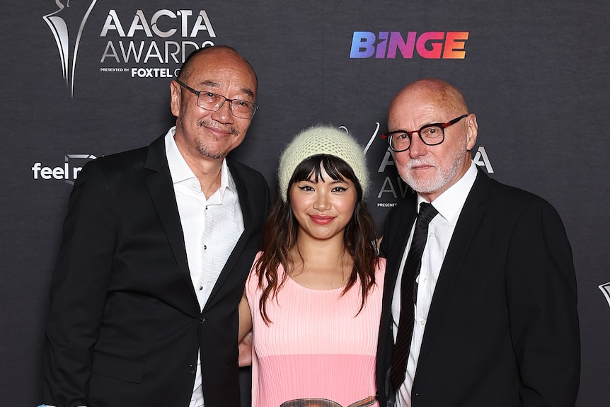 Asian-Australian man with glasses and a warm smile stands next to Asian-Australian woman in pink dress and white-bearded man.