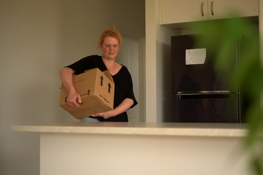 A woman with strawberry blonde hair is carrying a brown cardboard moving box into a kitchen.