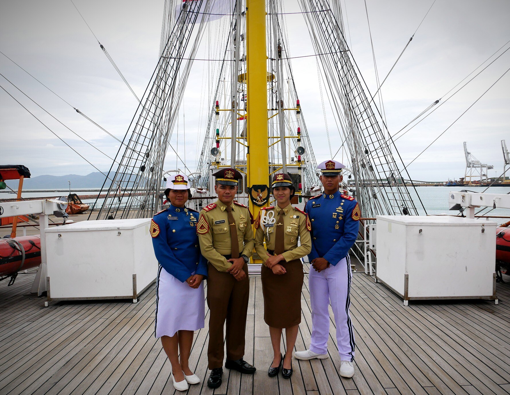 Four cadets in blue and green naval officer uniforms stand on deck of a sail ship