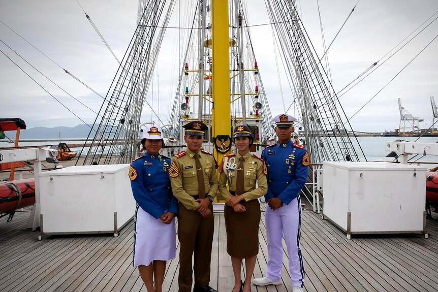 Four cadets in blue and green naval officer uniforms stand on deck of a sail ship