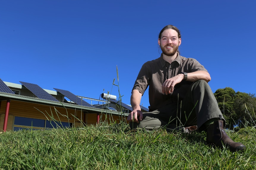Guy Stewart sits on the grass in front of building with solar panels