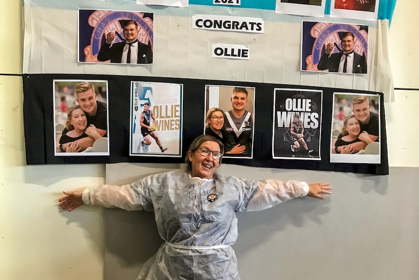 Woman wearing protective medical clothing and glasses smiles with arms stretched in front of wall of pictures of her son