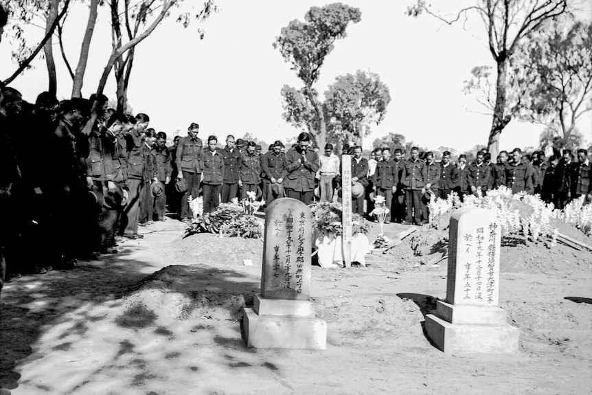 Dozens of men in uniform stand around graves. One man is standing in front of the grave with hands clasped in prayer.