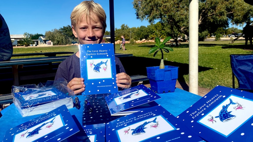 Smiling boy, blonde hair, sits behind a table covered with books in a park, trees, grass. Holds blue book with fish up. 