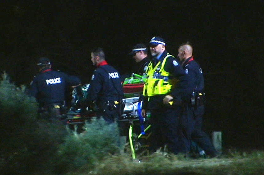 Police and paramedics accompany a woman on a stretcher at night in bushland.