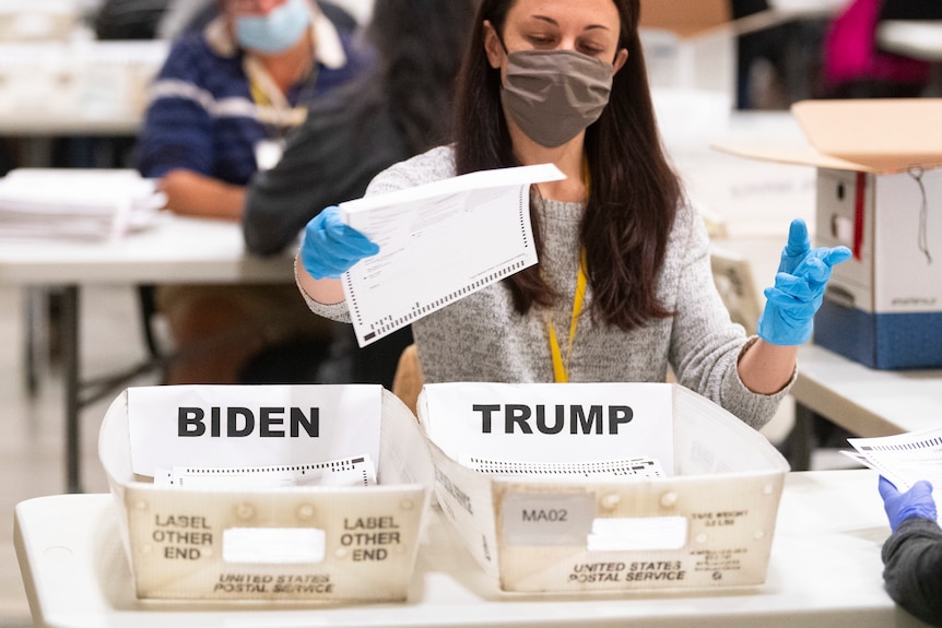 A woman in blue rubber gloves goes to place a paper ballot in either a Trump or Biden box.