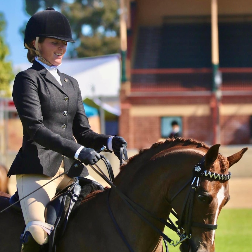 A female para-equestrian rider wears a jacket and helmet and is riding a horse in competition, smiling.