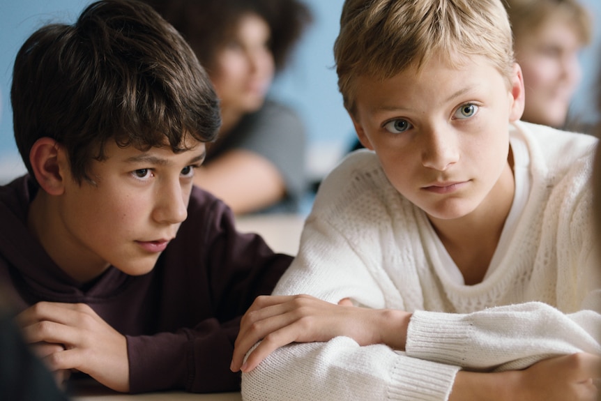 Two 13-year-old boys, one brown-haired the other blond, sitting close together as if in class, listening to someone.
