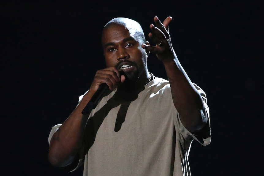Kanye West gestures at the camera with one hand, holding a microphone in the other.