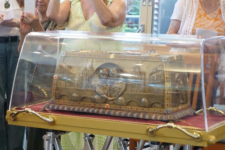 People gathered around a glass case housing an intricate reliquie box made out a bronze like material