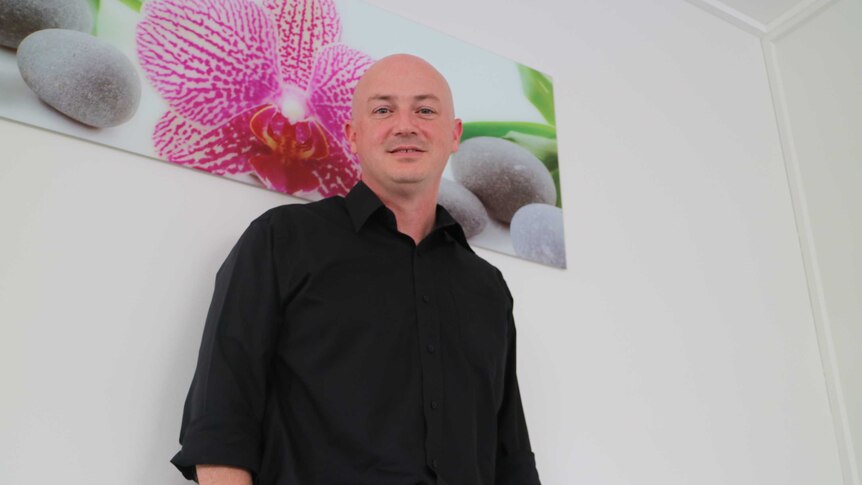 A bald man in a black shirt stands in front of a painting of a pink flower