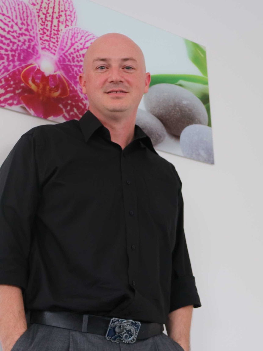 A bald man in a black shirt stands in front of a painting of a pink flower