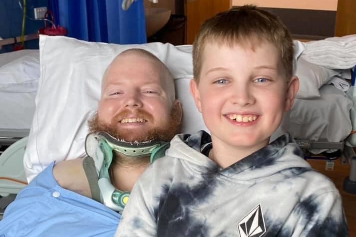 A man lying in a hospital bed in a neck brace is smiling after a visit from his young son.