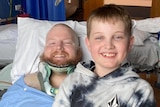 A man lying in a hospital bed in a neck brace is smiling after a visit from his young son.