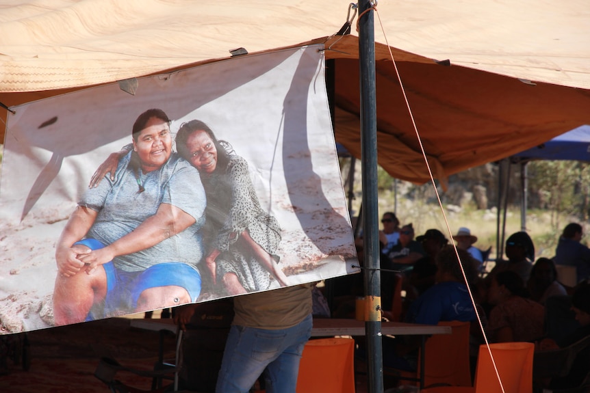A poster hanging from a large tent of a younger lady smiling in the arms of an older lady.