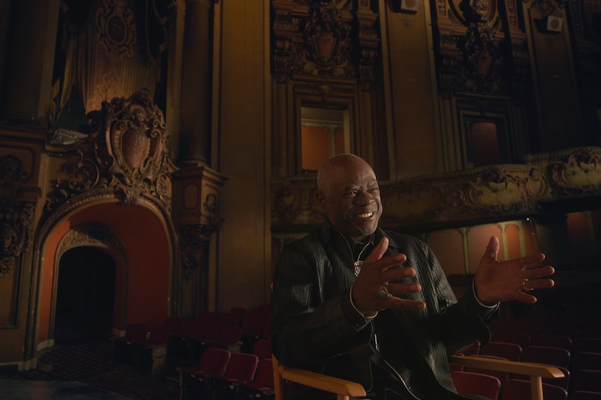 Bald middle-aged black man sits in chair wearing all black inside an ornately decorated theatre auditorium.