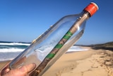 Hand holding a scotch bottle with a tightly-rolled letter inside, beach in background.