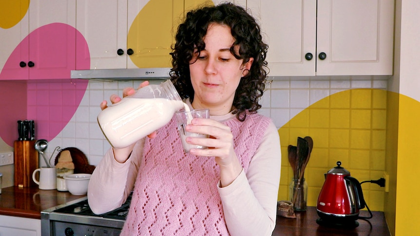 Phoebe Thorburn looks at a glass as she pours non-dairy milk into it in her kitchen, with cut out pink and yellow blobs.
