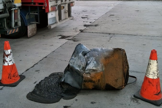 A fire-damaged barrel of chemicals on the ground outside a factory between two cones.