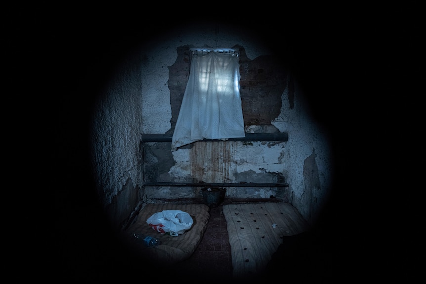 Two tattered, dirty-looking mattresses lie on the floor of a dark, dank room.