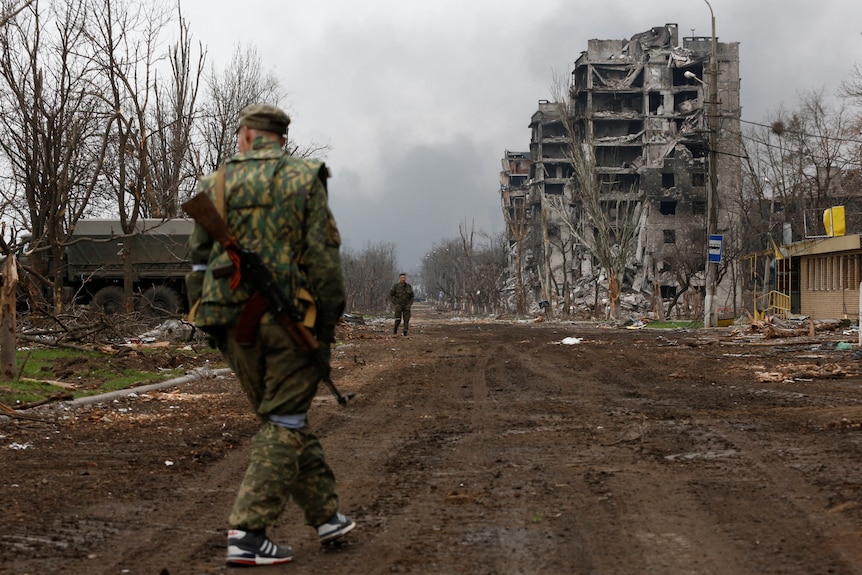 A soldier walks along a road with a destroyed building in the background.