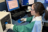 A medical technician insert a cartridge into a small machine. Computers are on in the background.
