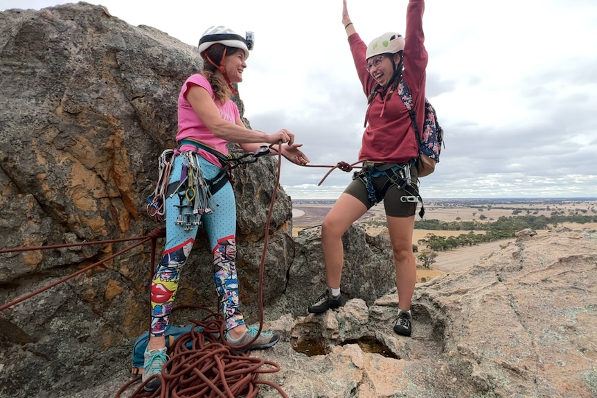 a girl in a red hoodie and brown shorts and white helmet with her hands up as she climbed to the top with a climbing instructor