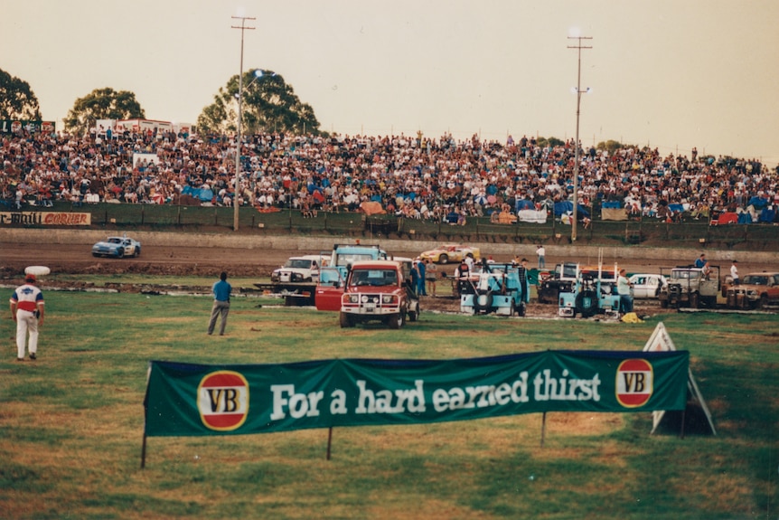 A scattering of trucks and cars sit on a green oval, surrounding by cars speeding along a dirt track. A packed crowd looks on.