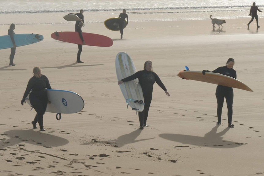 A group of 7 female surfers walking away from the waves, carrying their boards