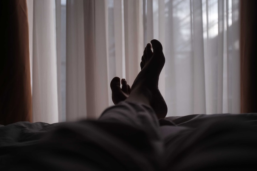 The view of feet in bed, in a dark room, with feint light through sheer curtains in the background.
