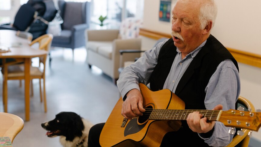 An older man sits on a chair holding a guitar next to a black and white dog