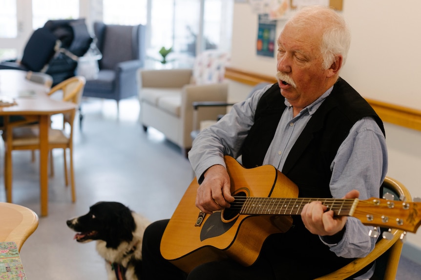An older man sits on a chair holding a guitar next to a black and white dog