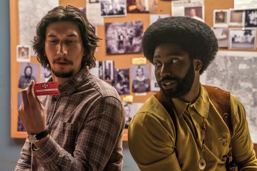 Adam Driver in plaid shirt looking at KKK membership card, sitting next to John David Washington with afro and mustard shirt.