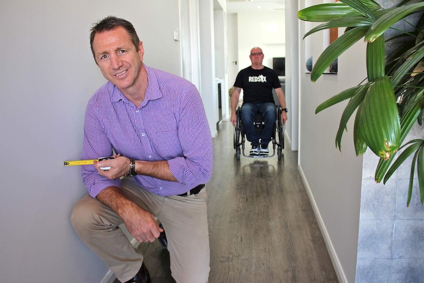 A man holds a builder's measuring tape up to a hallway wall, behind him in the hallway is a man in a wheelchair.