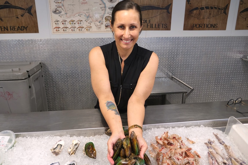 a woman stands in seafood resturant holding oysters in her hands smiling at the camera.