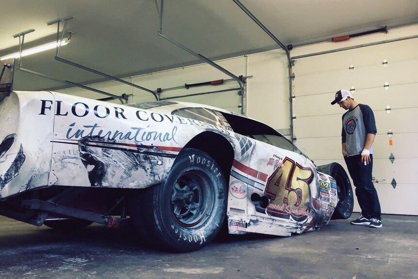 A bent and charred NASCAR vehicle is looked at by its injured driver.