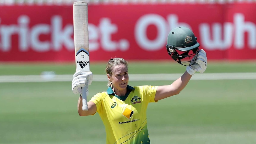 Ellyse Perry raises her bat and helmet as she stands in the middle of a cricket field.