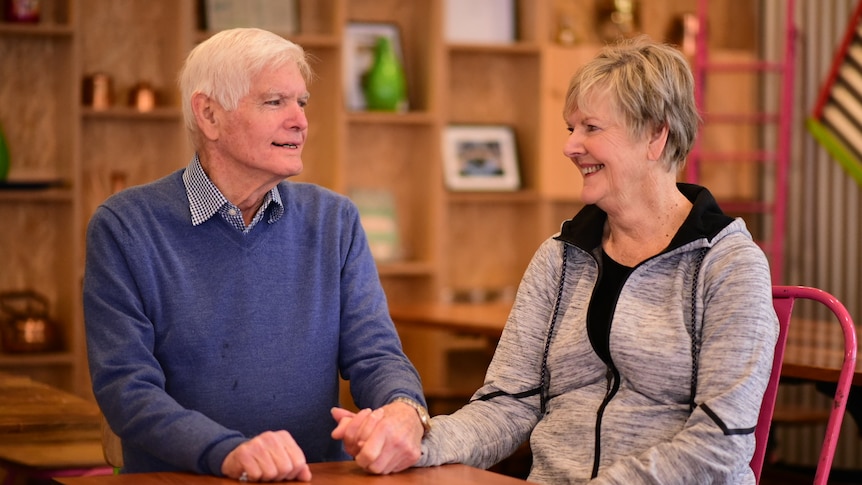 A man and woman sit at a red table in a cafe, holding hands on the table. They are looking at each other and smiling