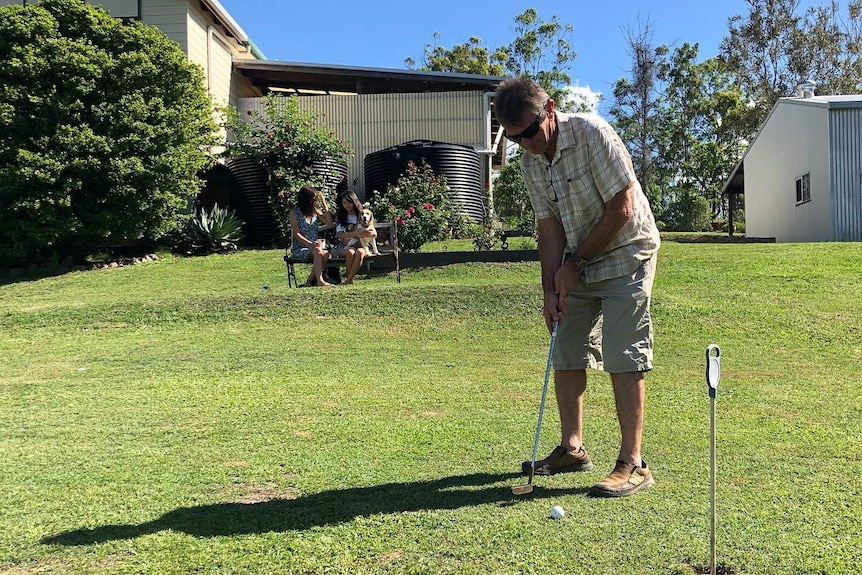 Mr Binney swings a golf club in his back yard with his wife and daughter looking on.