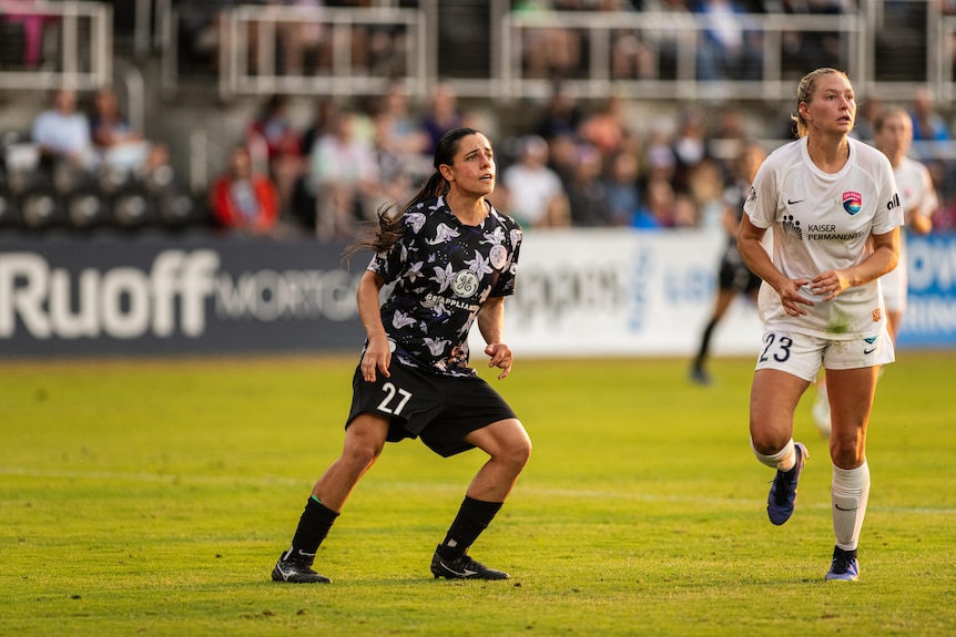 El futbolista australiano Alex Chidiac posa en el campo con Racing Louisville FC para NWSL mientras un jugador vestido de blanco lo salta.