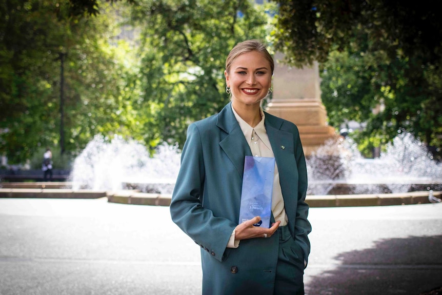 A young woman stands in front of a fountain holding an award