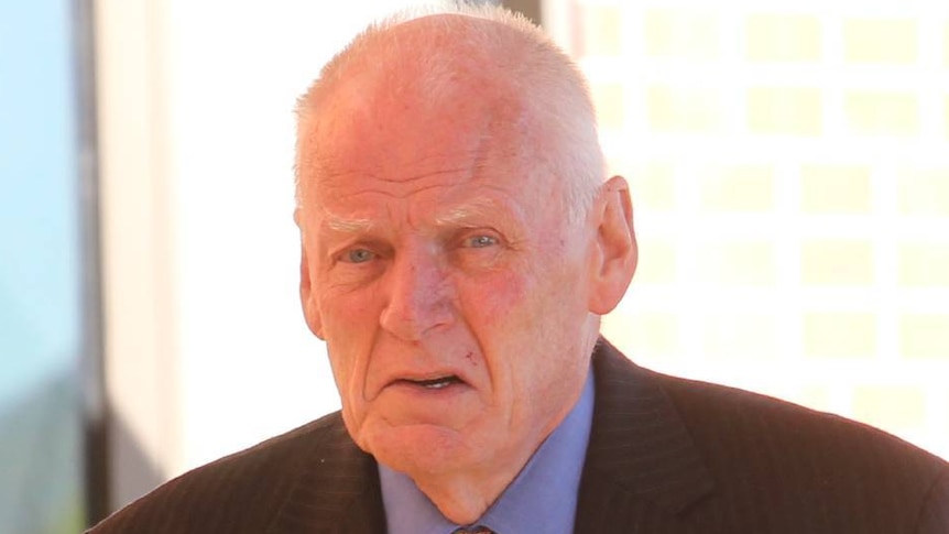 An elderly looking man with thin white hair in a black suit looks at the camera as he walks past.